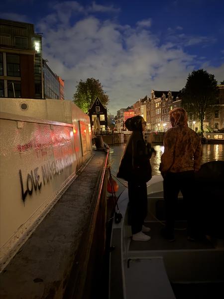Two people stand on a canal boat, looking out at the illuminated historic buildings lining the water in Oudeschans, Uilenburg, Amsterdam